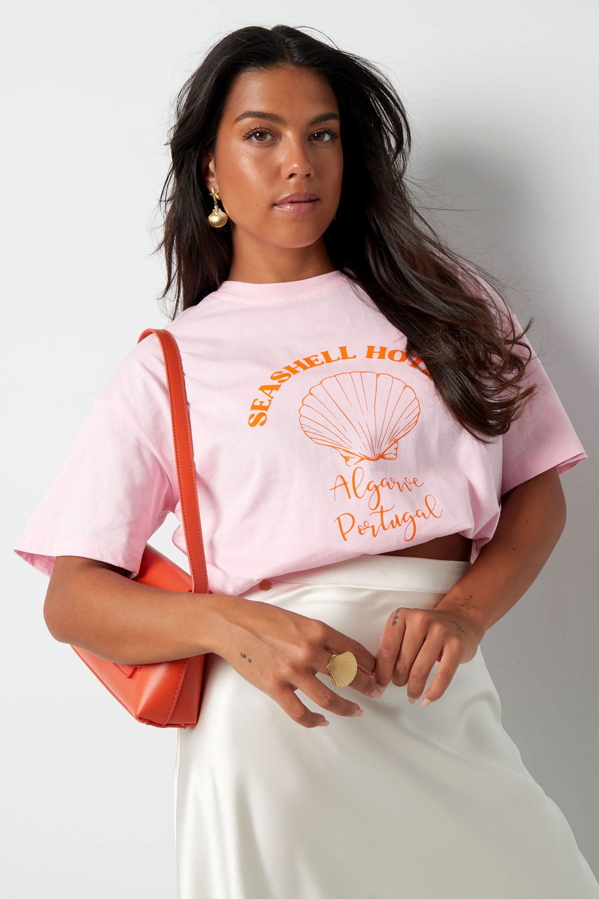 T-Shirt "Seashell Hotel" Rosa – stylisches Baumwoll-T-Shirt mit Muschel-Aufdruck und orangefarbener Schrift auf der Vorderseite.